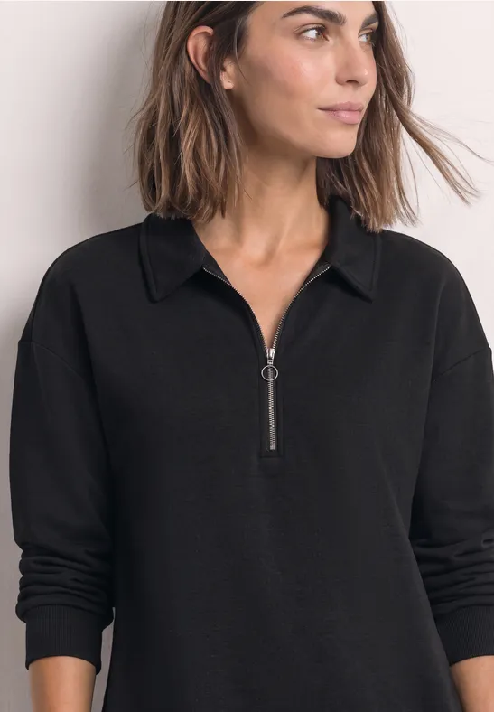 Frau in schwarzem Langarmshirt mit Reißverschluss und Kragen, schulterlanges braunes Haar, frontalaufnahme.
