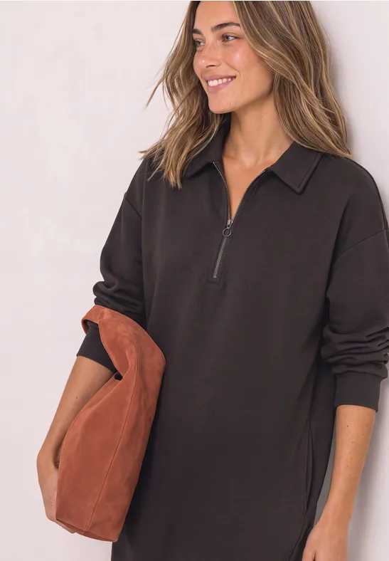 Femme en robe noire à manches longues zippée, souriante, tenant un grand sac marron.