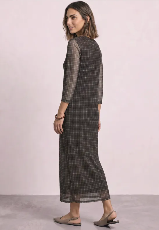 Frau in langem kariertem Maxikleid mit 3/4-Ärmeln und flachen Sandalen, seitlich stehend.