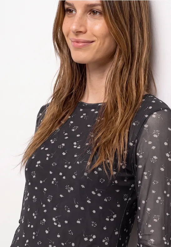 Frau in schwarzem, leicht transparentem Oberteil mit kleinem weißen Blumenmuster und dezentem Lächeln.