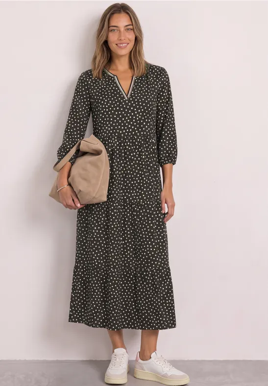 Frau in langem schwarzen Polka-Dot-Kleid mit beigem Beutel und weißen Sneakern vor hellem Hintergrund.