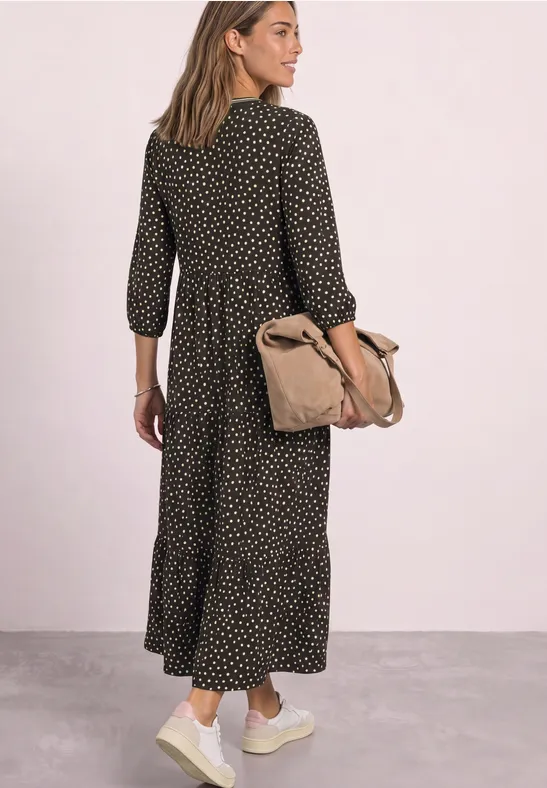 Frau von hinten in schwarzem gepunktetem Midi-Kleid, trägt Sneaker und eine beige Umhängetasche vor rosafarbenem Hintergrund.