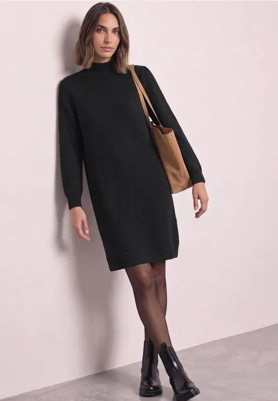 Frau in schwarzem Pulloverkleid mit Strumpfhose, schwarzen Boots und brauner Umhängetasche vor hellem Hintergrund.