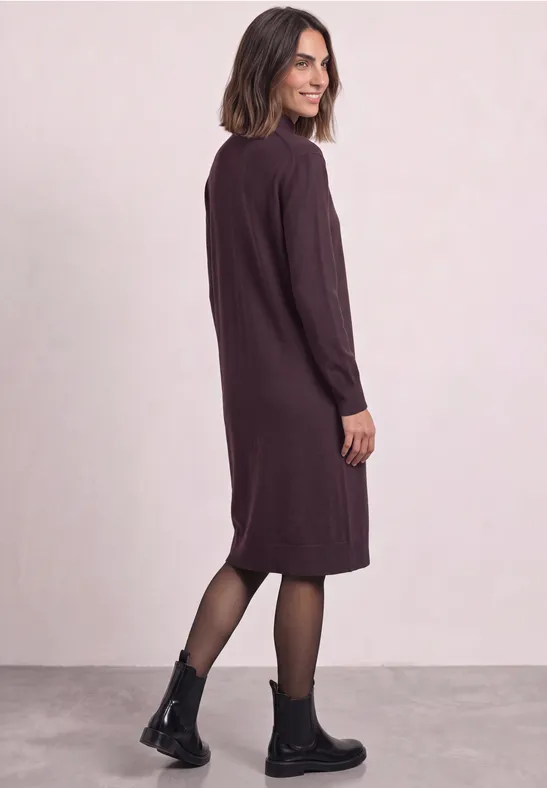 Frau in knielangem, langärmeligen, dunkelviolettem Kleid mit schwarzen Boots und Strumpfhose, seitlich stehend.