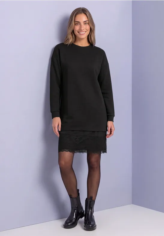 Frau in schwarzem Strickkleid mit Spitzenbesatz, schwarzen Strumpfhosen und Stiefeletten vor hellvioletter Wand.