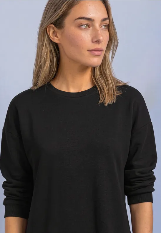 Frau trägt schlichten schwarzen Pullover, blickt zur Seite, vor neutralem Hintergrund.
