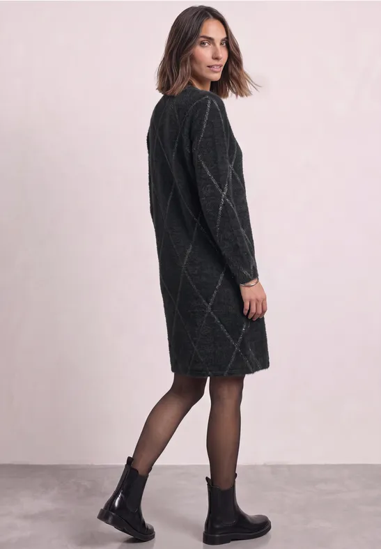 Frau im schwarzen, diamantgemusterten Strickkleid, blickt über die Schulter, trägt schwarze Stiefel und Strumpfhose.