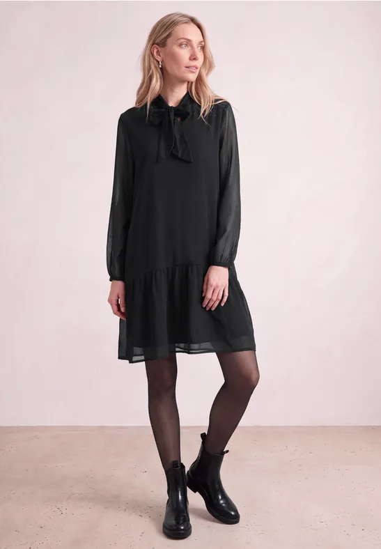 Eine Frau vor hellem Hintergrund in schwarzem Kurzkleid mit Schleife, schwarzen Strumpfhosen und Stiefeletten.