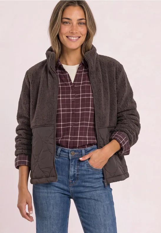 Lächelnde Frau trägt braune Fleecejacke über kariertem Hemd und blaue Jeans.