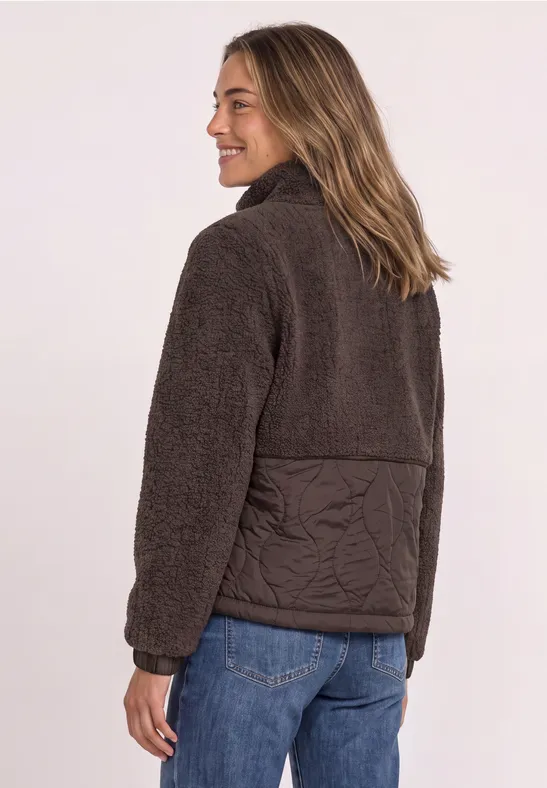Frau in brauner Fleecejacke mit Steppung auf Rückenpartie und Jeans, Blick zur Seite.