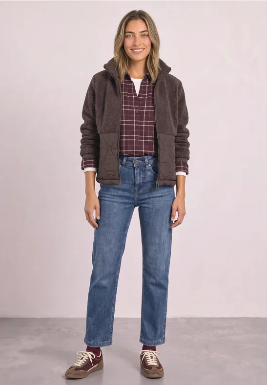 Frau trägt kariertes Hemd, braune Jacke, gerade Jeans und Sneaker, steht vor rosa Hintergrund.
