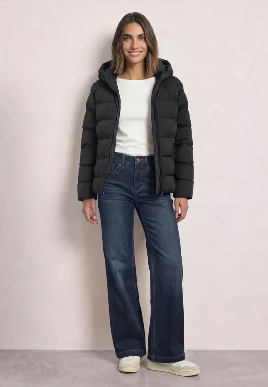 Frau im schwarzen Steppmantel, weißem Pullover und weitem Jeans-Look steht lächelnd da.