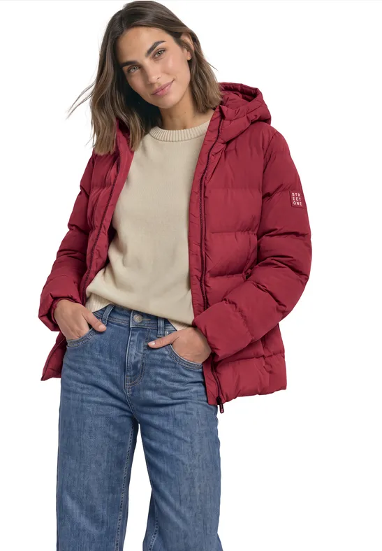 Vrouw in een rode donsjas, beige trui en blauwe spijkerbroek tegen een witte achtergrond.