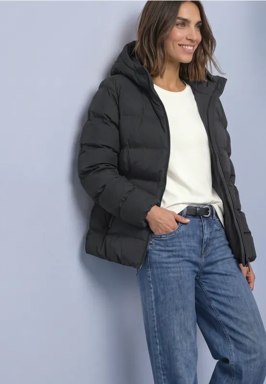 Femme appuyée contre un mur, portant une doudoune noire, un pull blanc et un jean bleu, souriante.