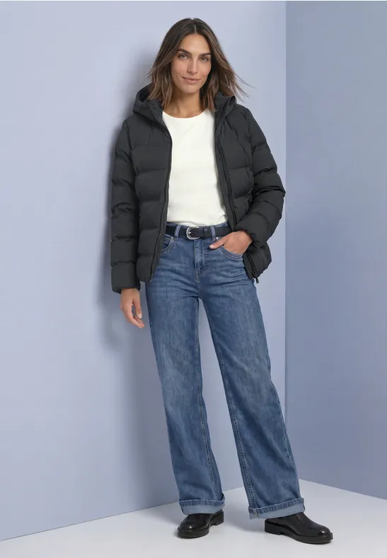 Femme en doudoune noire, t-shirt blanc, jean bleu et chaussures noires devant un mur bleu clair.
