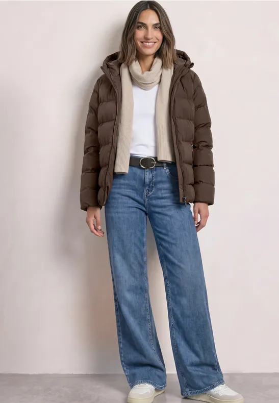 Femme souriante en doudoune marron, pull blanc, jean, écharpe et ceinture.
