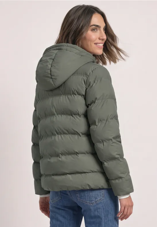 Femme portant une doudoune kaki à capuche et un jean, regarde par-dessus l’épaule avec un sourire.