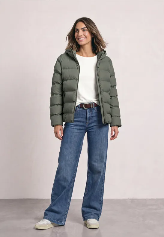 Femme souriante portant doudoune verte, t-shirt blanc et jeans bleu, posant devant un fond neutre.