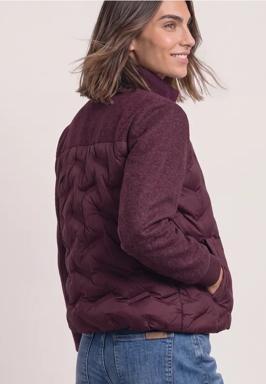 Frau mit schulterlangem Haar trägt eine warme, weinrote Steppjacke und Jeans, blickt lächelnd zur Seite.