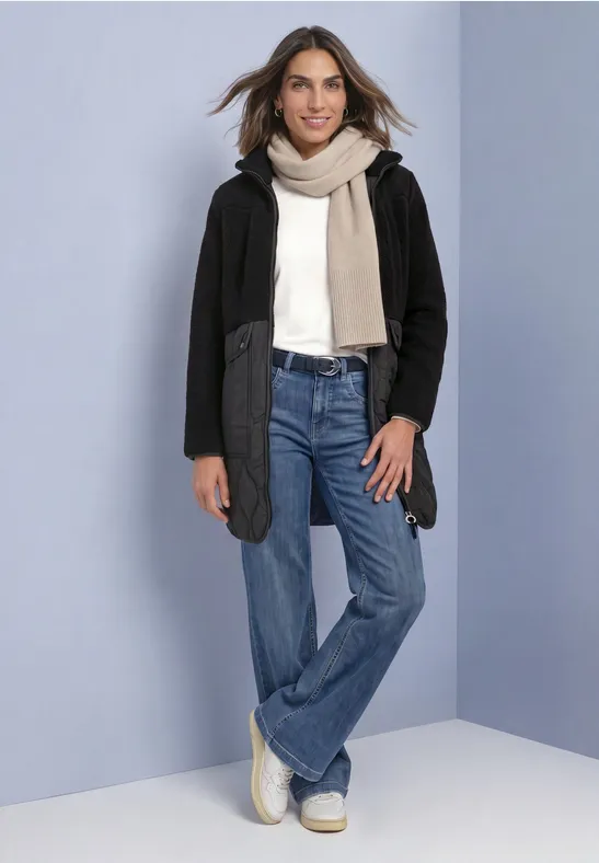 Eine Frau in Jeans, hellem Pullover, Schal und schwarzer Jacke lächelt vor hellblauem Hintergrund.