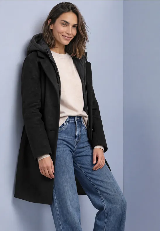 Femme portant un manteau noir, pull clair et jeans bleus, souriante devant un fond neutre.