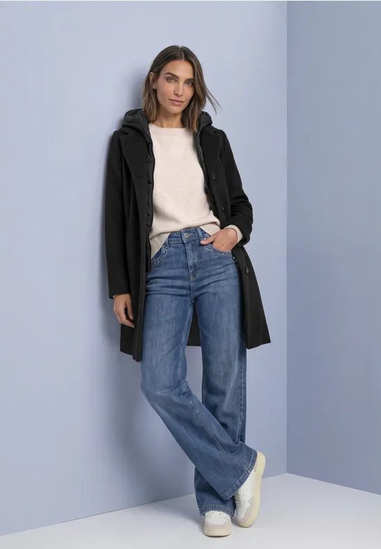 Femme appuyée contre un mur bleu clair, manteau noir, pull blanc, jeans bleu et baskets blanches.