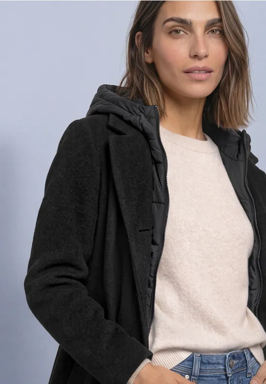 Femme en manteau à capuche noir doublé, pull beige et jean, posant devant un fond simple.
