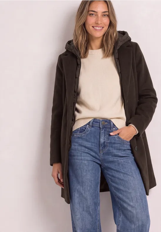 Junge Frau trägt braunen Parka über hellem Pullover und blauen Jeans, posiert lässig.