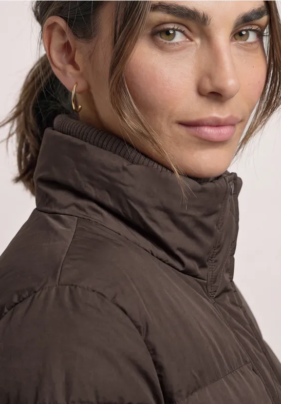 Frau mit brauner Steppjacke und hohem Kragen, dezentes Make-up, dunkle Haare, schaut in die Kamera.