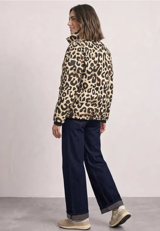 Vrouw met luipaardprintjas, wijde jeans en sneakers voor een lichte achtergrond.
