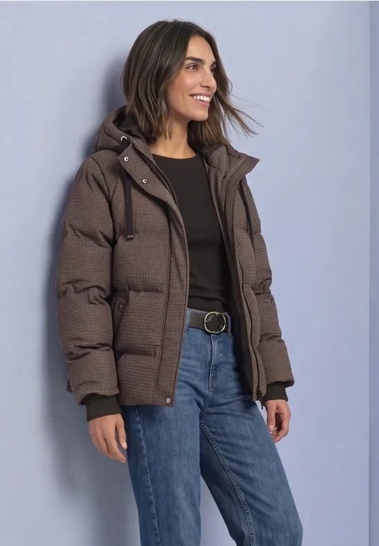 Frau mit brauner Daunenjacke, schwarzem Shirt und blauen Jeans lächelt seitlich.