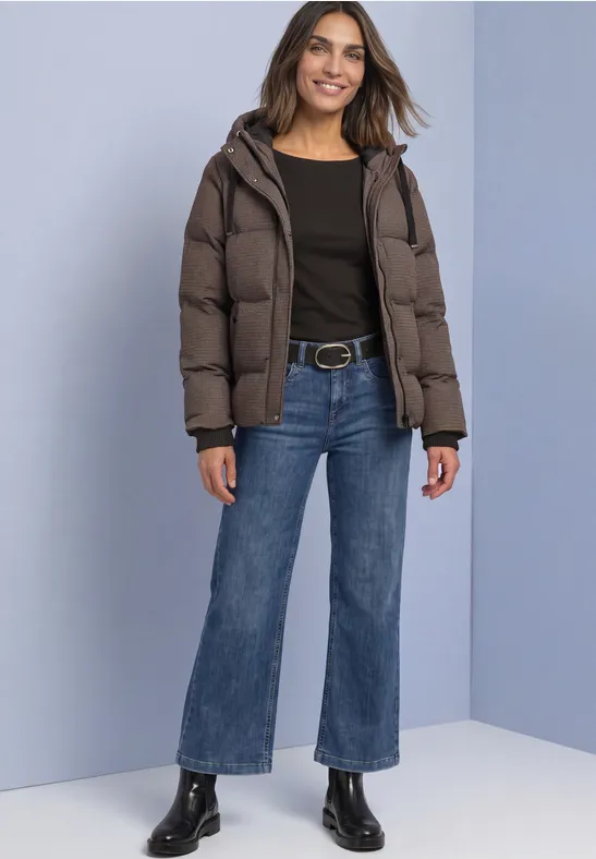 Eine Frau posiert lächelnd in einer Daunenjacke, schwarzem Top, blauen Jeans und schwarzen Stiefeletten.