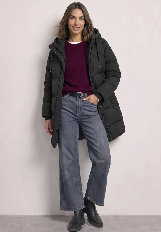 Eine lachende Frau in schwarzer Steppjacke, weinrotem Pullover, weitem Jeans und schwarzen Stiefeletten.