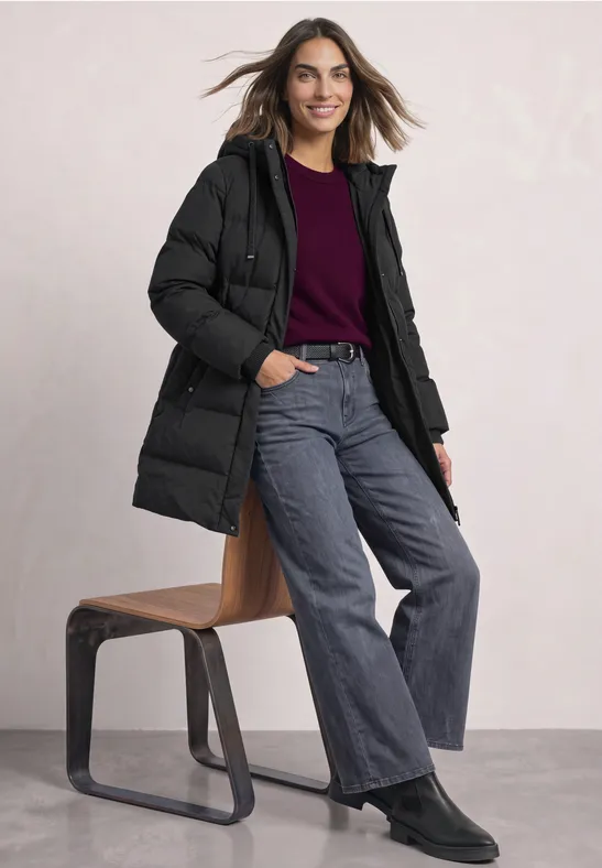 Frau in schwarzer Daunenjacke, weinrotem Pullover, weitem Jeans und schwarzen Stiefeletten lehnt lächelnd an einem Hocker.