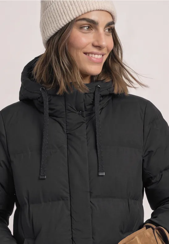Frau mit beige Strickmütze und schwarzer Steppjacke, lächelnd im Winterstyling.