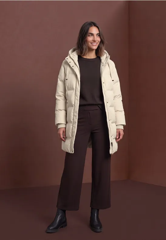 Vrouw in beige donsjas, bruine outfit en zwarte enkellaarsjes, poseert voor een bruine achtergrond.