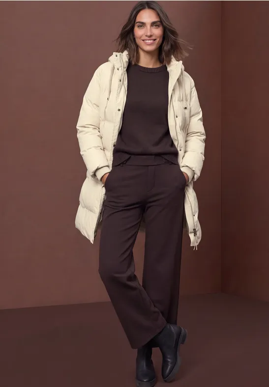 Vrouw glimlachend in lange beige donsjas, bruin outfit en zwarte laarzen, voor een bruine achtergrond.