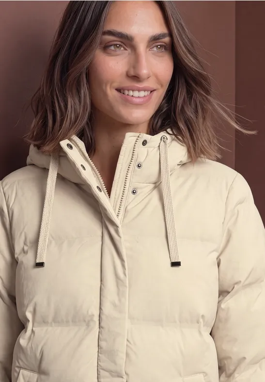 Vrouw met halflang bruin haar lacht, draagt een lichtbeige gewatteerde jas met capuchon en trekkoorden.