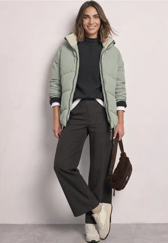 Frau in grüner Jacke mit Teddyfutter, schwarzem Pullover, weiter Hose und weißen Sneakern, braune Tasche.