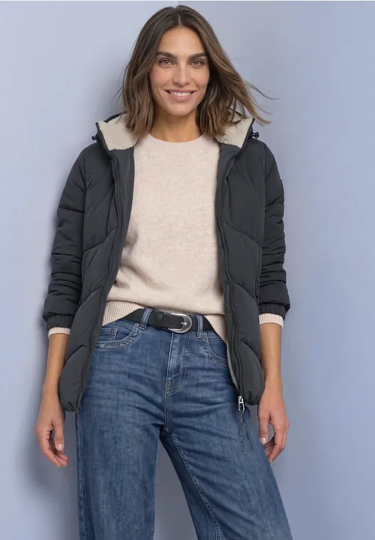 Frau in schwarzer Steppjacke mit Kapuze, beigem Pullover und blauen Jeans, lächelnd.