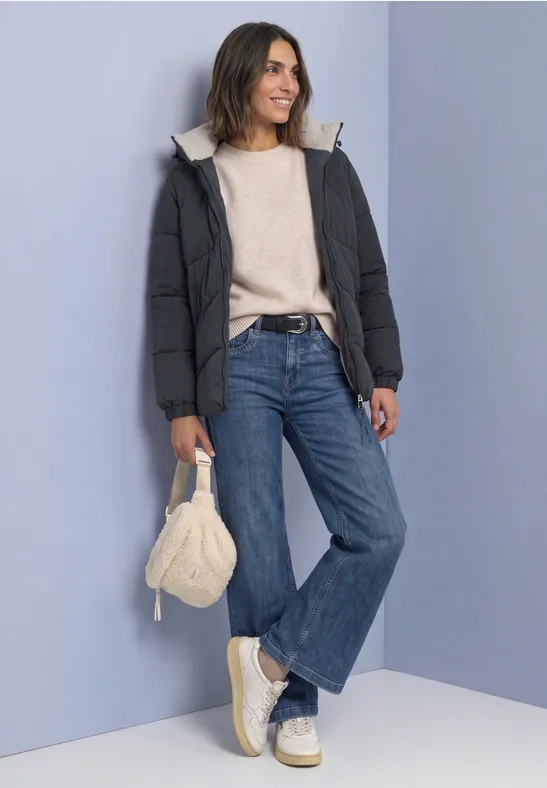 Frau in Steppjacke, Pullover, weiter Jeans, Plateauschuhen und Stricktasche vor lila Wand