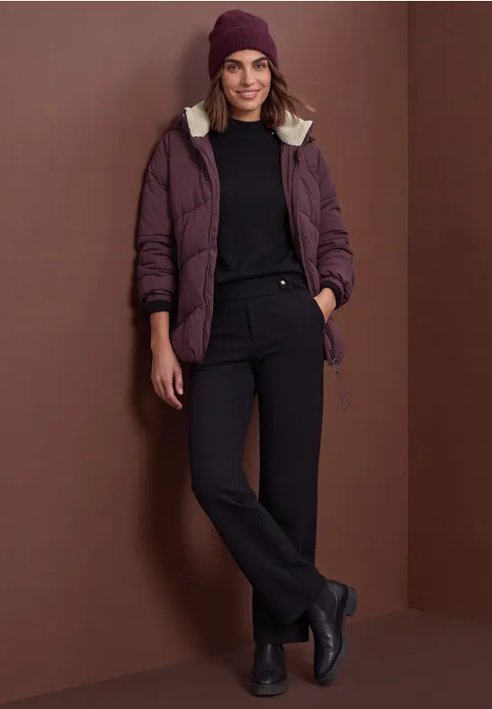 Frau in beerenfarbener Jacke, schwarzem Rollkragenpullover, schwarzer Hose, Boots & Mütze vor braunem Hintergrund.