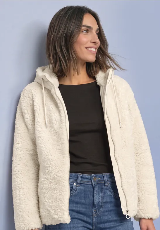 Frau in weißer Teddy-Fleecejacke mit Kapuze, schwarzem Shirt und Jeans, lächelnd zur Seite blickend.