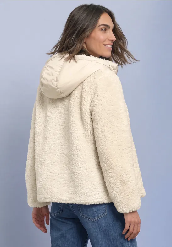 Frau in cremefarbener Teddyjacke mit Kapuze und Jeans, leicht seitlich von hinten.