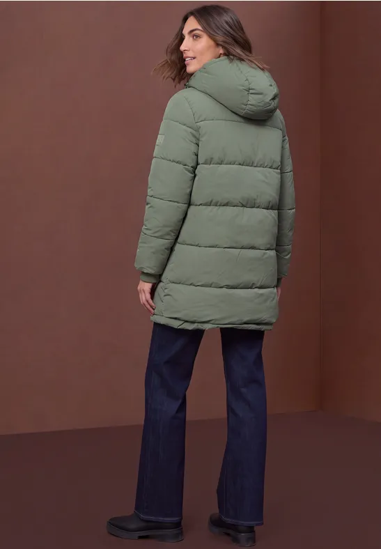 Frau in grüner Steppjacke, blauen Jeans und schwarzen Schuhen, steht vor braunem Hintergrund und blickt zurück.