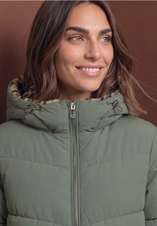 Eine lächelnde Frau trägt eine grüne, gesteppte Jacke mit Kapuze vor braunem Hintergrund.