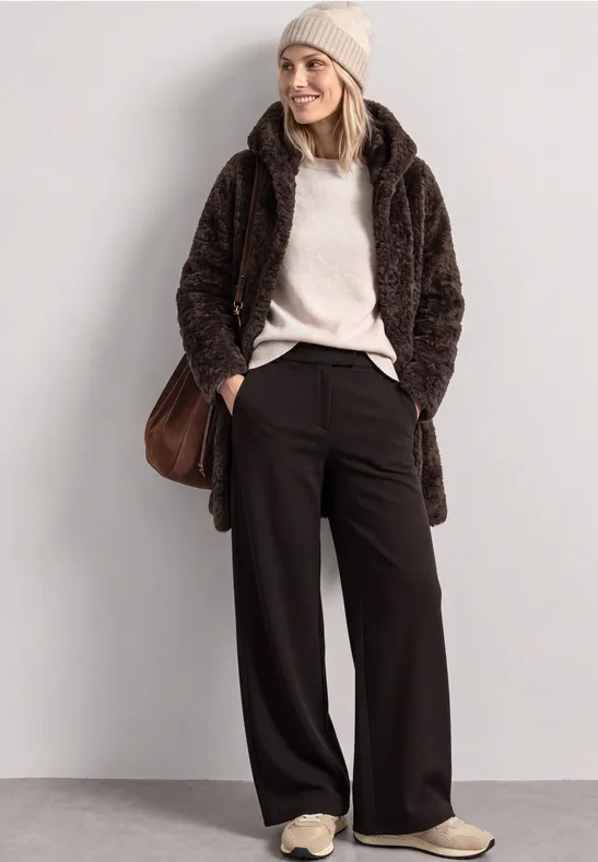 Frau trägt braunen Teddy-Mantel, beigen Pullover, schwarze weite Hose, Beanie und Sneakers.