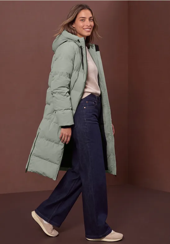 Frau in langem mintgrünem Daunenmantel, heller Bluse und weiten Jeans, vor braunem Hintergrund.