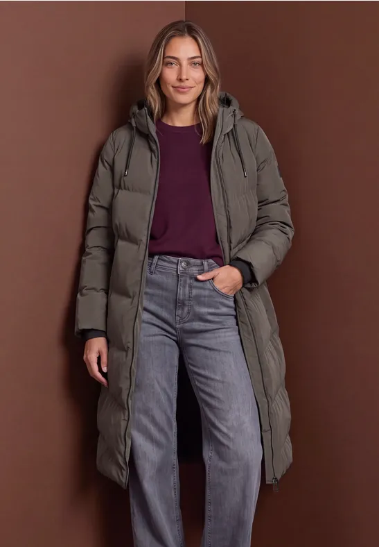 Frau in langem Steppmantel mit Pullover und Jeans vor braunem Hintergrund.