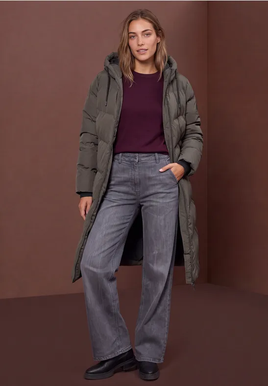 Frau in langem Daunenmantel, weinrotem Oberteil und weiten grauen Jeans vor braunem Hintergrund.